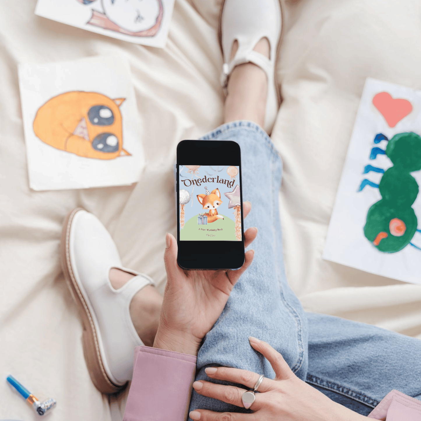 Person holding a phone with a cartoon fox on a screen, surrounded by children's drawings on a light-colored surface. Onederland Childrens Picture Book.